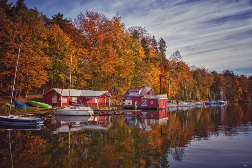 Fotografia łodzi oraz domków przy jeziorze, w tle jesienny las.