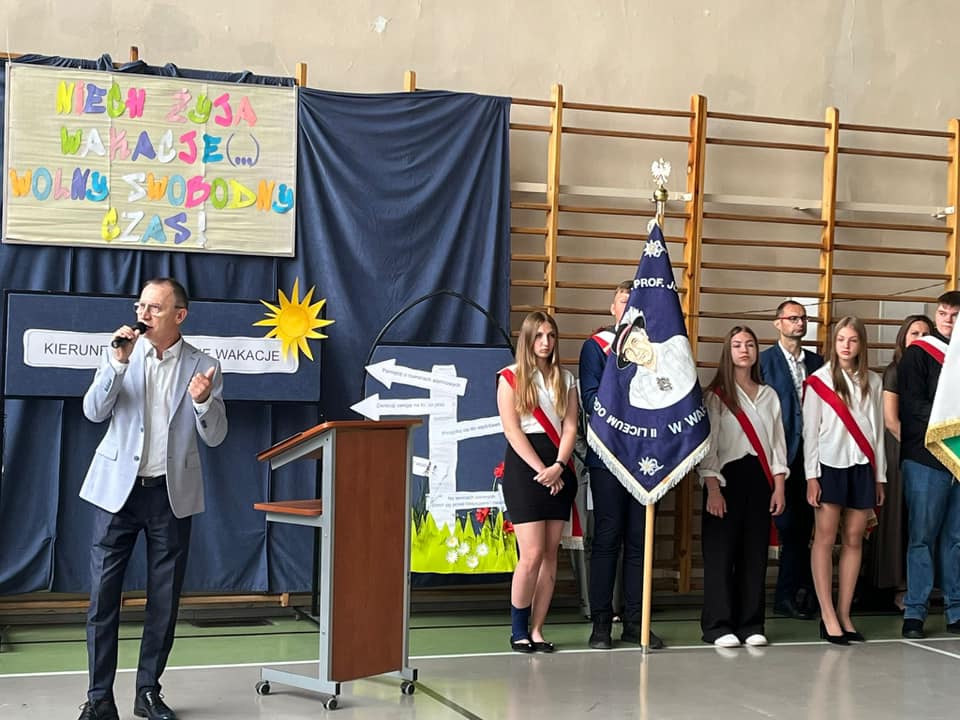Zdjęcie przemawiającego dyrektora oraz uczniowie stojący ze sztandarem szkoły.